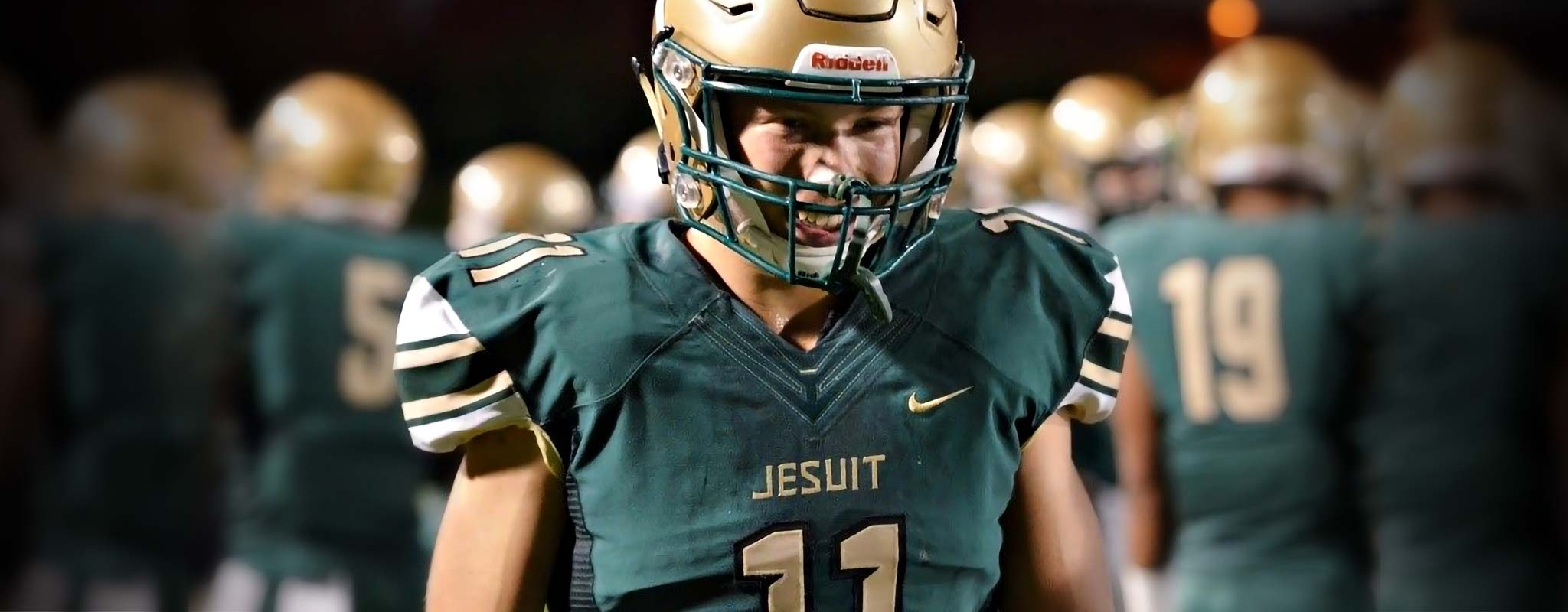 A Jesuit high school football player in front of his teammates, garbed in colors of gold and green.