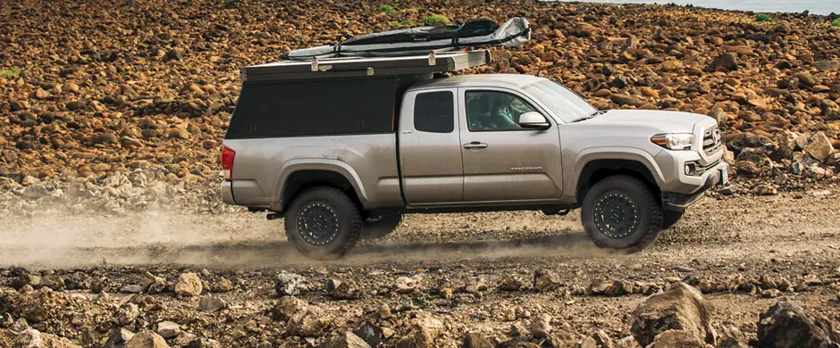 Small silver truck with camper driving over rough gravel road.